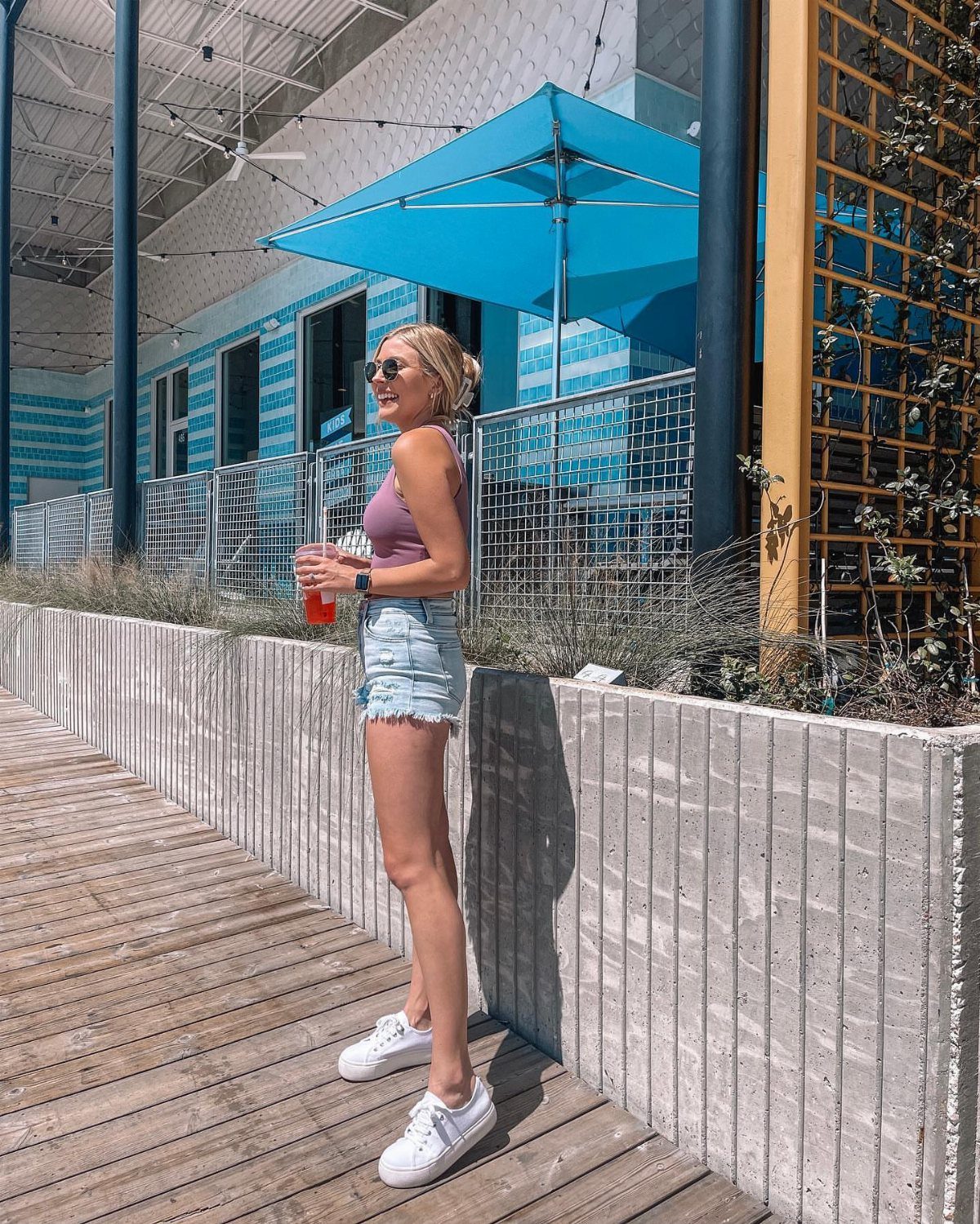 A smiling young woman wearing a purple crop top and denim shorts standing on a wooden walkway.