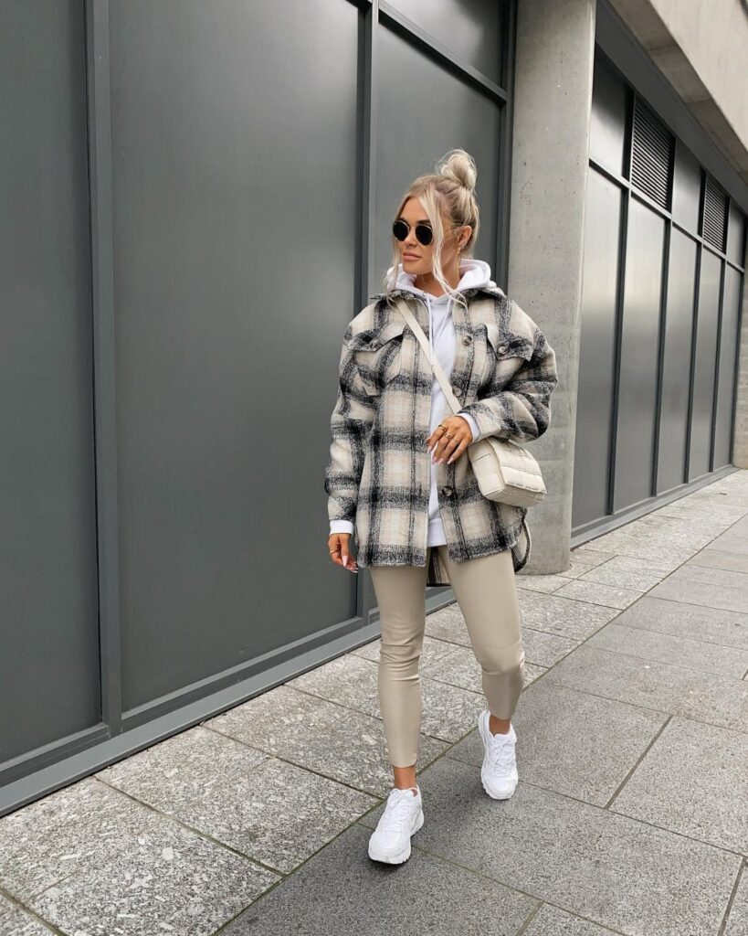 A woman wearing a plaid shacket, white hoodie, beige leggings, and white sneakers outdoors.