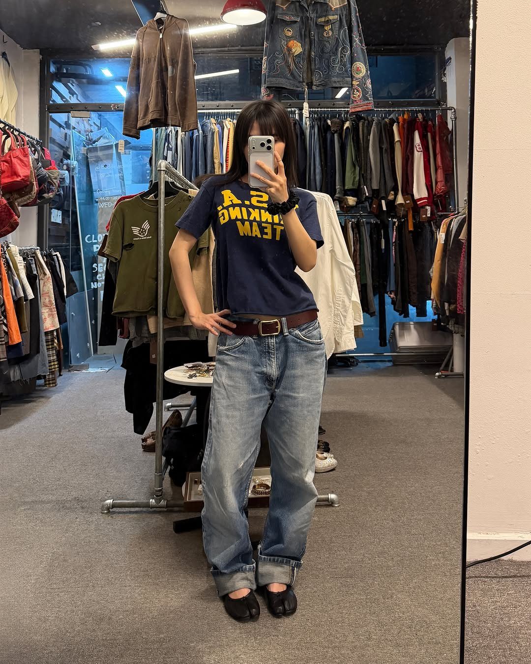 Person wearing a navy graphic t-shirt and baggy blue jeans posing in a clothing store mirror.