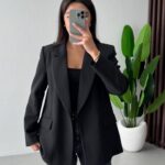 A woman posing in a black blazer, black top, and dark jeans while taking a selfie.