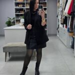 A woman in an all-black outfit with leather skirt and black boots posing in a closet.
