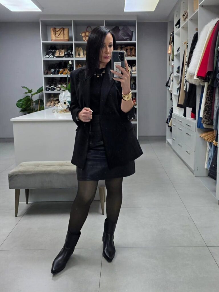 A woman in an all-black outfit with leather skirt and black boots posing in a closet.