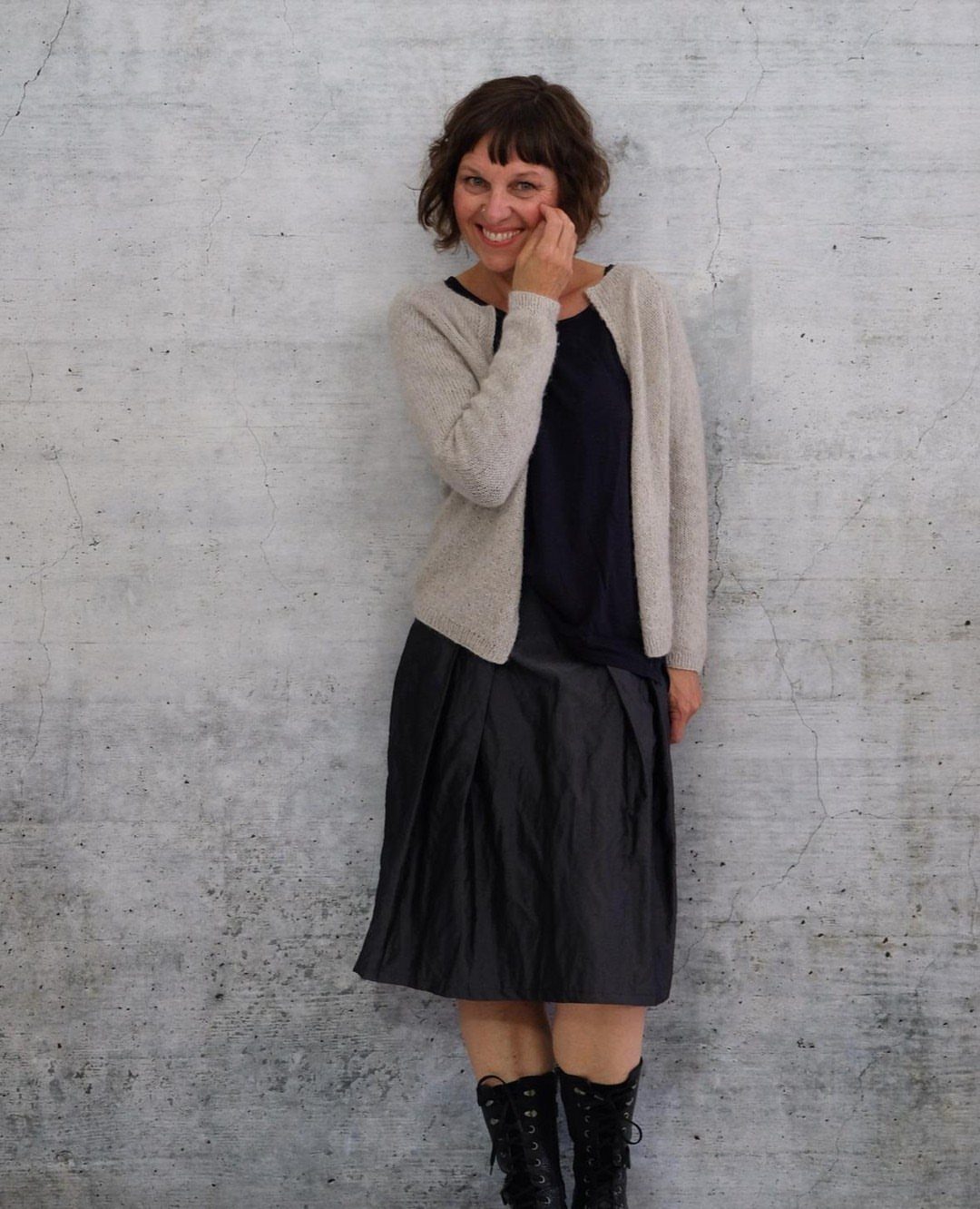 A smiling woman wearing a beige cardigan, navy top, black midi skirt, and black boots.