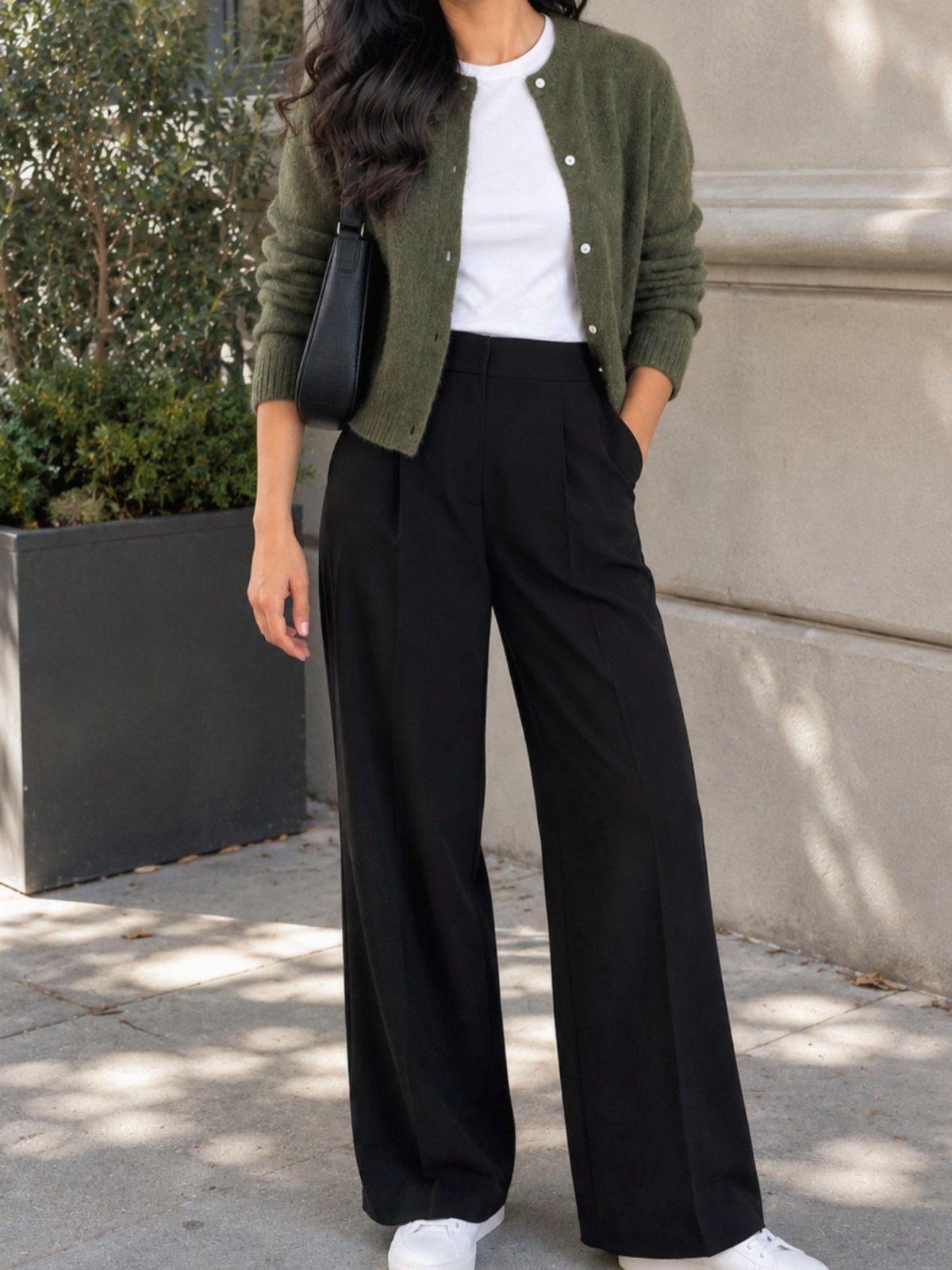 A person wearing an olive green cardigan, white shirt, and black wide-leg trousers outdoors.