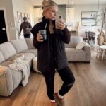 A blonde woman wearing a black puffer jacket and leggings taking a mirror selfie.