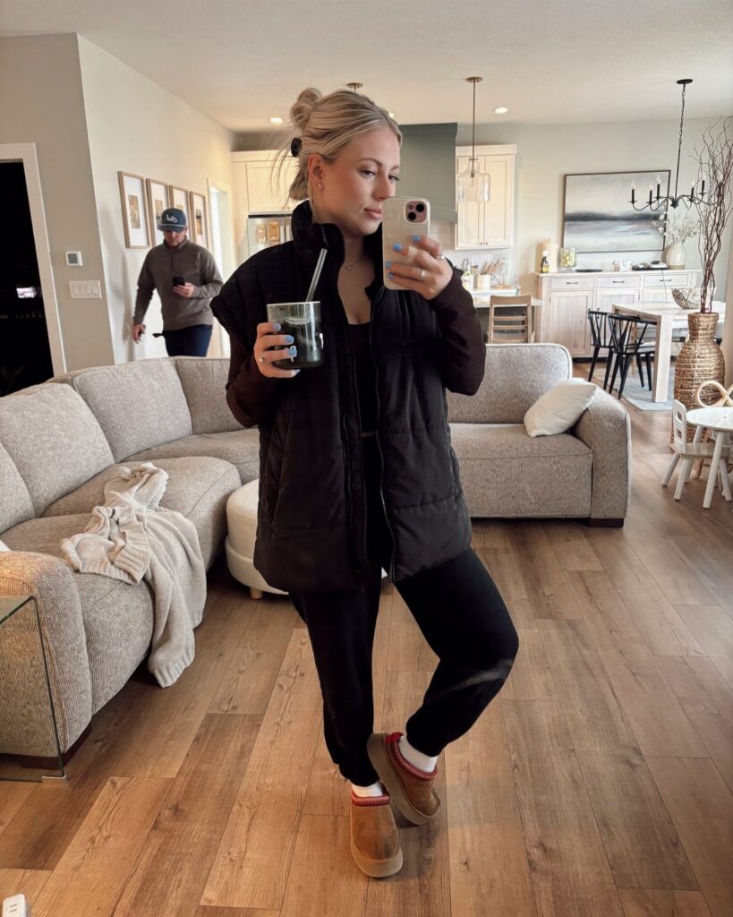 A blonde woman wearing a black puffer jacket and leggings taking a mirror selfie.