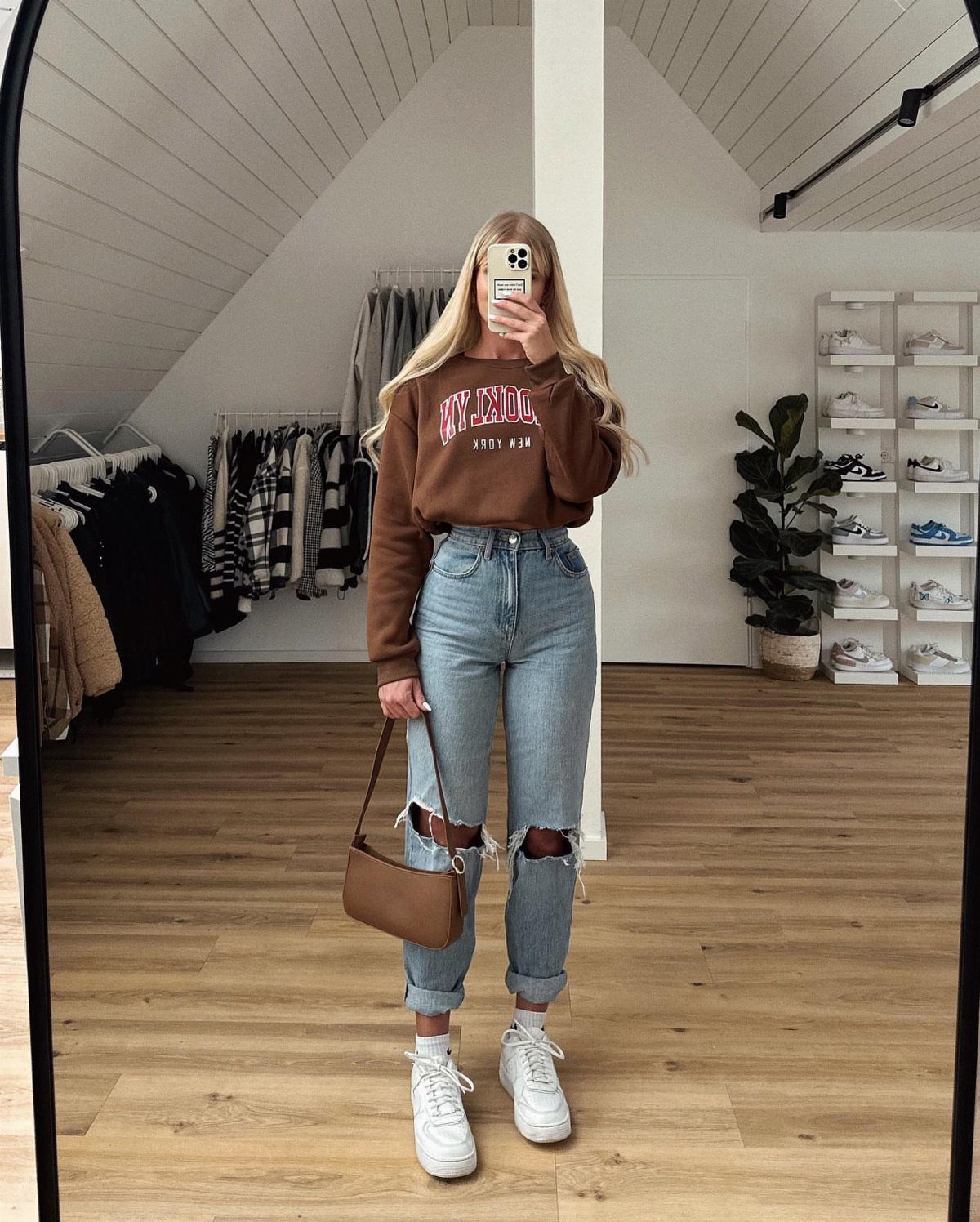 Woman in a brown sweatshirt and ripped blue jeans posing for a mirror selfie.