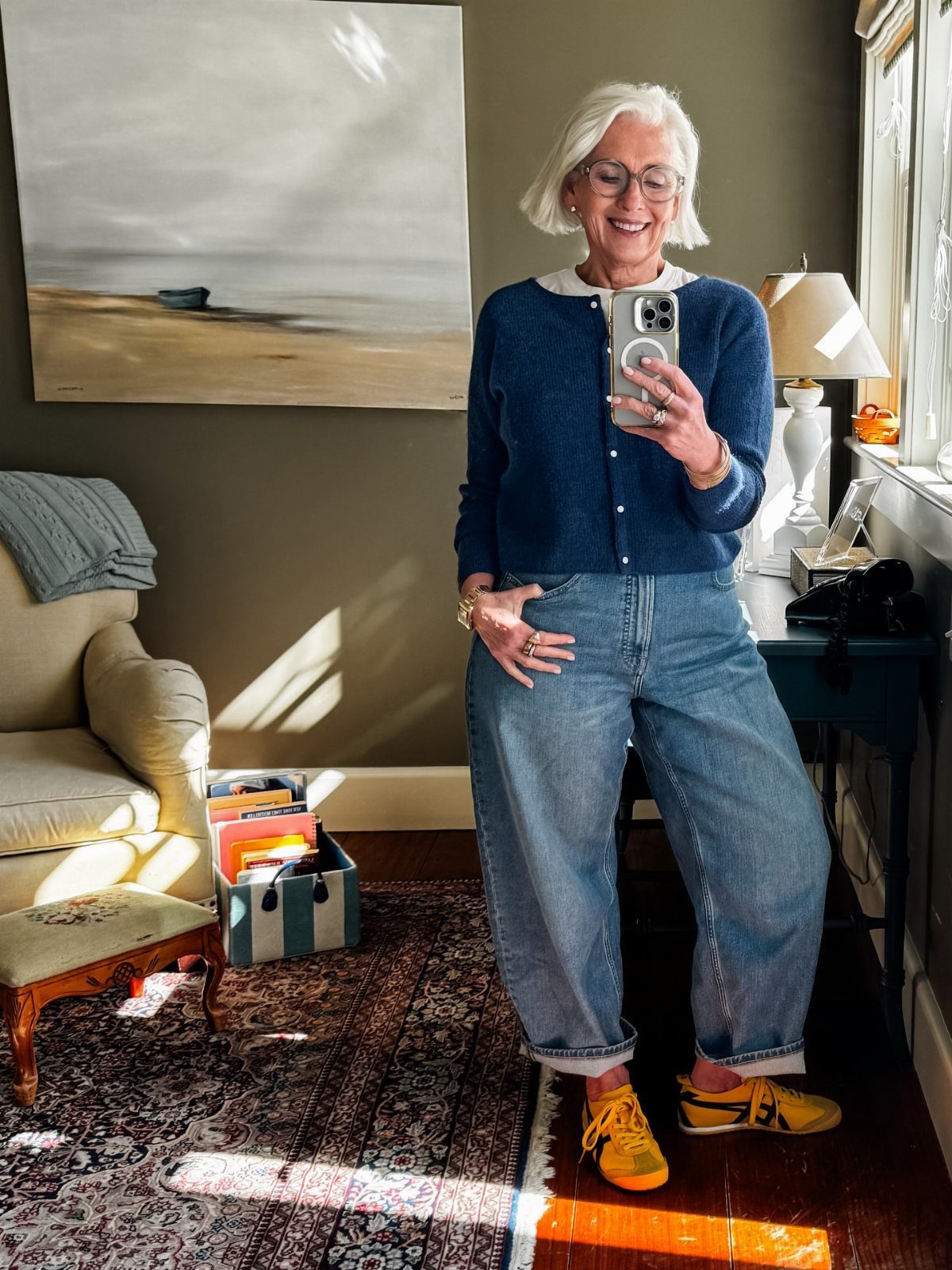 A smiling woman with white hair wearing a blue cardigan, baggy jeans, and yellow sneakers.