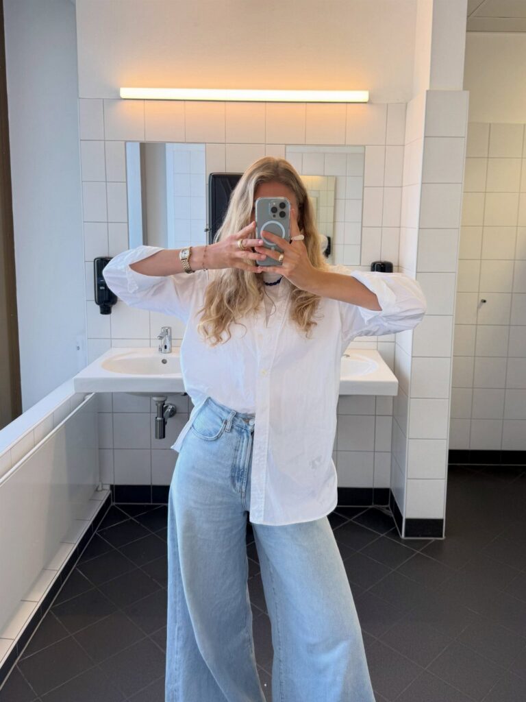Woman in a white button-down shirt and wide-leg blue jeans taking a mirror selfie.