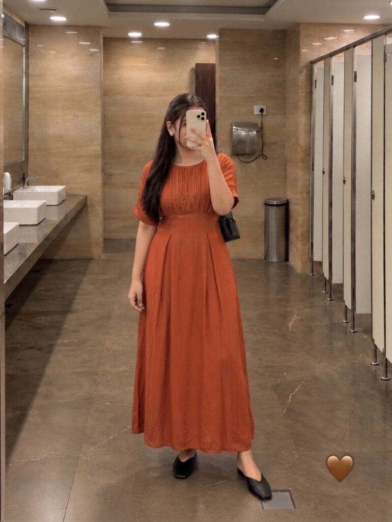 A woman in a long burnt orange pleated dress posing for a mirror selfie.