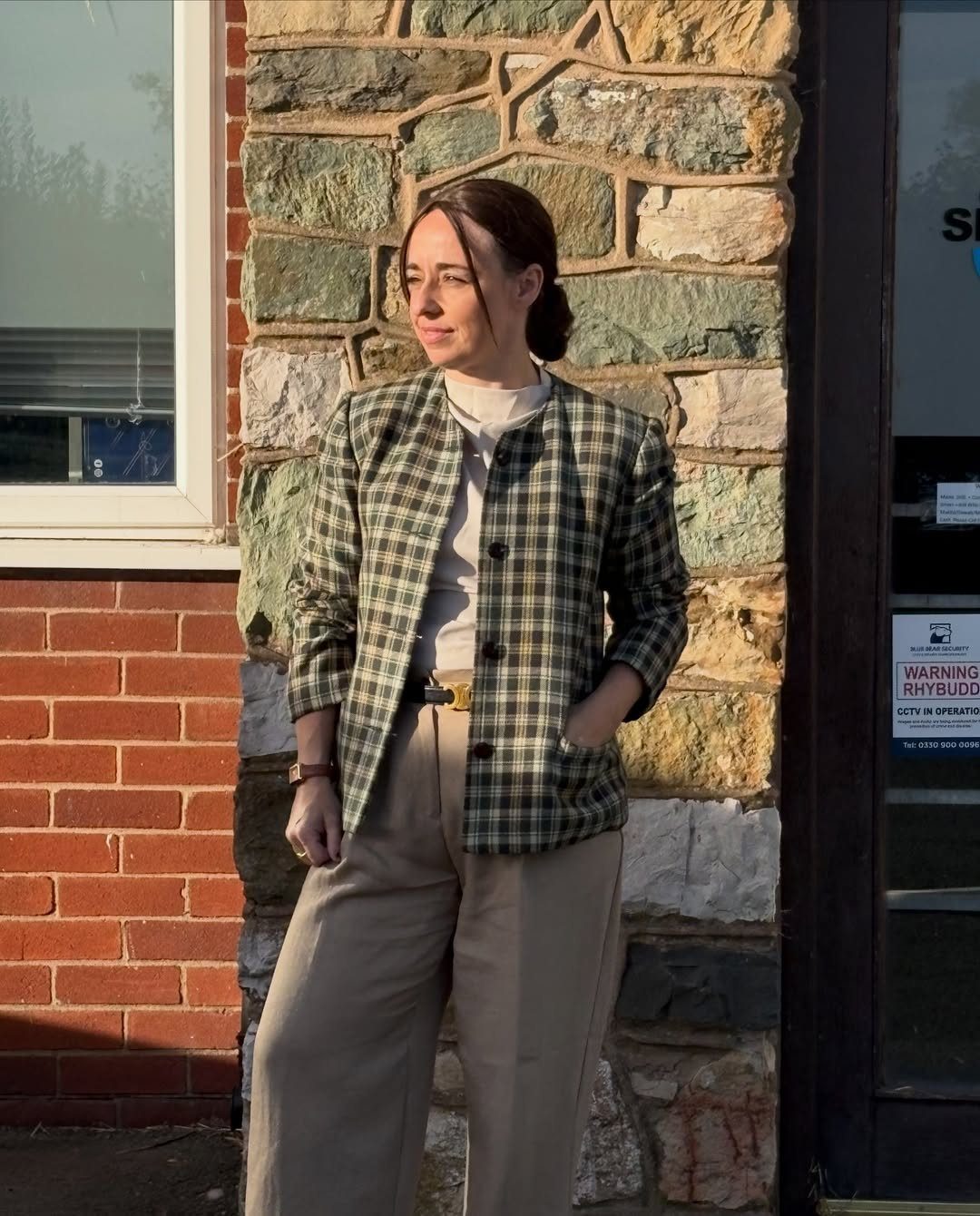 A woman wearing a green plaid blazer and beige trousers standing against a stone wall.