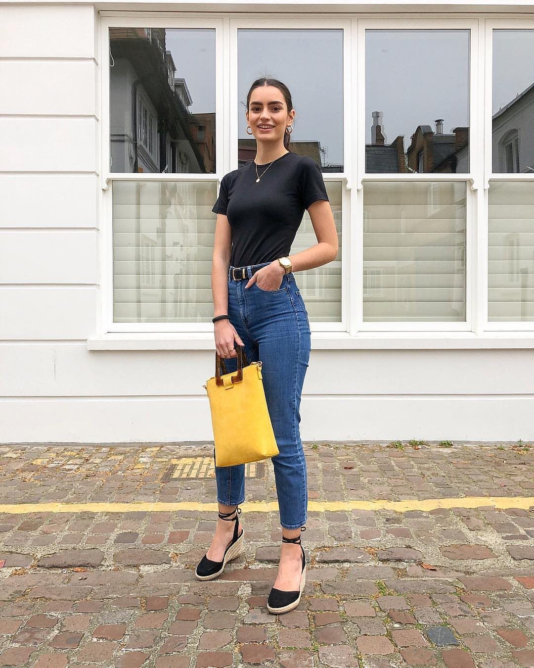 A woman in a black t-shirt and blue jeans holding a yellow handbag.