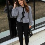 A woman wearing a checkered peplum blouse, black trousers, and black flats standing outdoors.