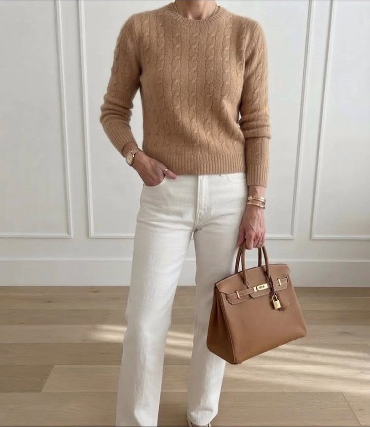 A person wearing a camel cable-knit sweater and white trousers while holding a tan handbag.