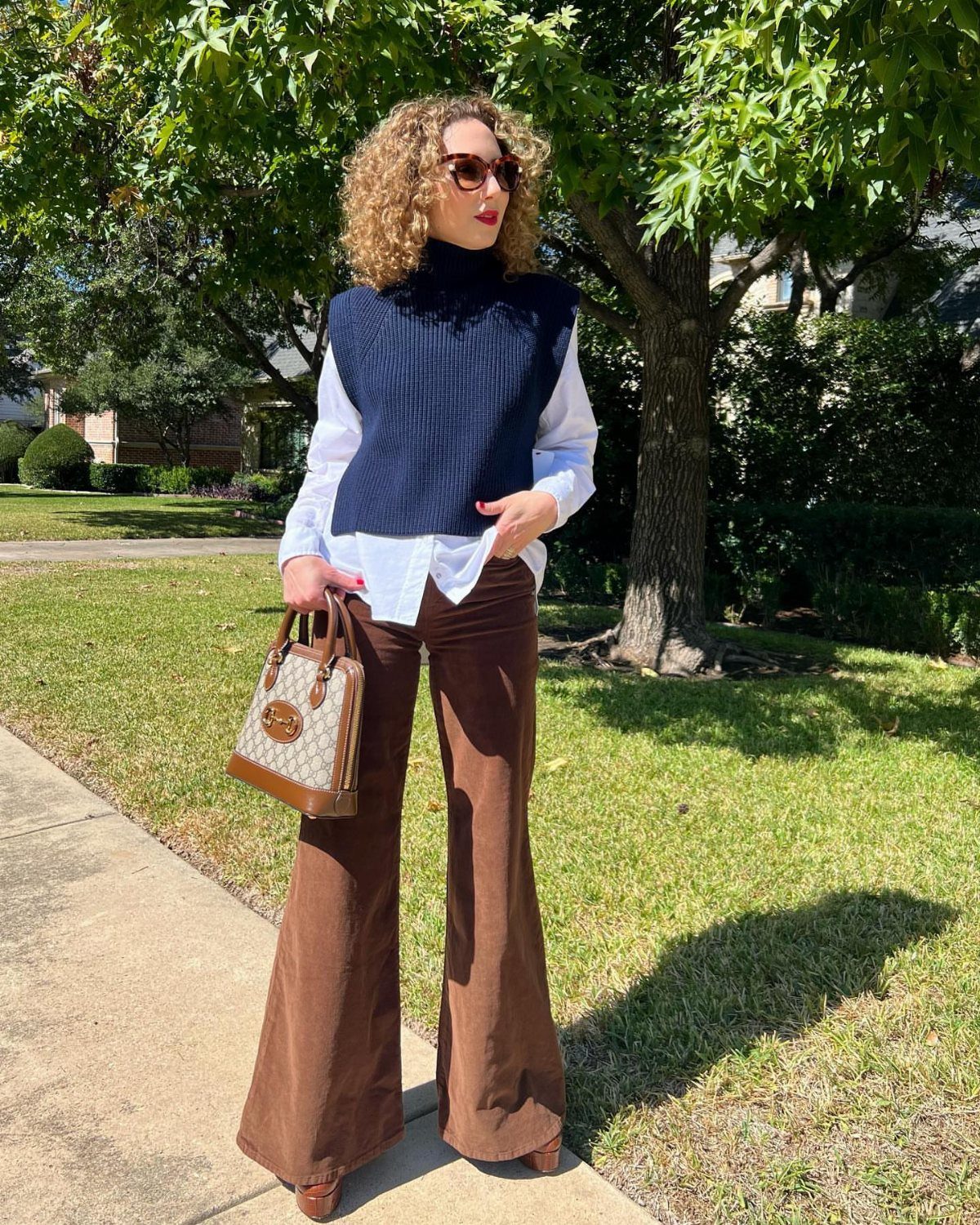 Woman wearing a navy cable knit sweater vest over a white shirt and brown trousers.