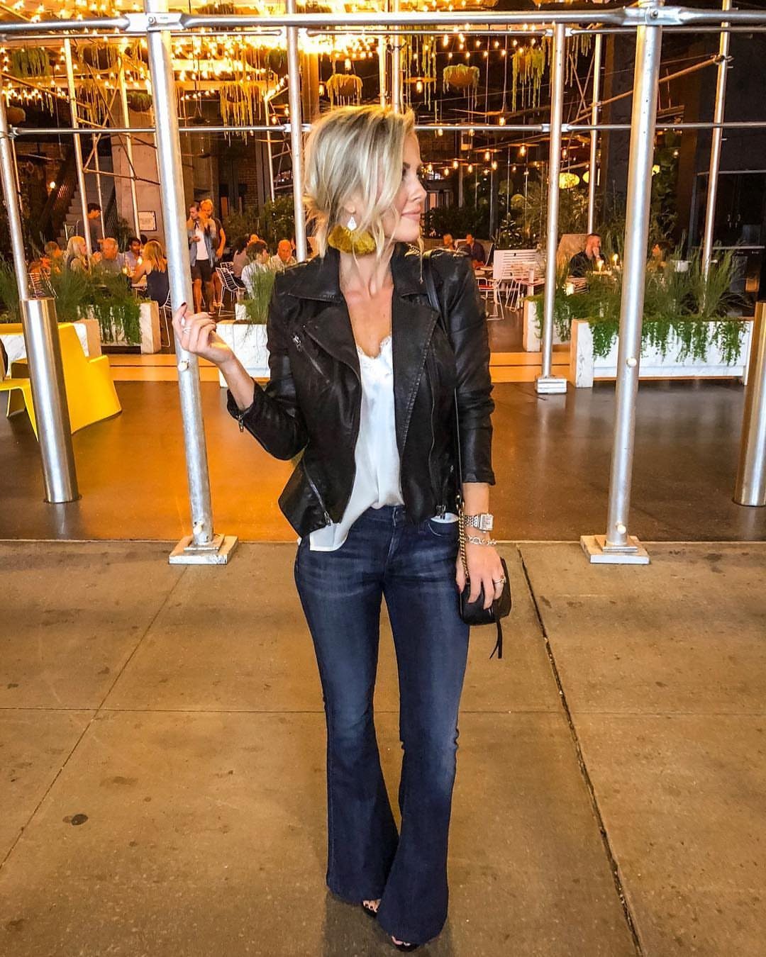 A blonde woman posing in a black leather jacket and dark wash flare jeans outdoors at night.