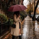 A woman in a tan trench coat walks along a rainy city sidewalk under a burgundy umbrella.