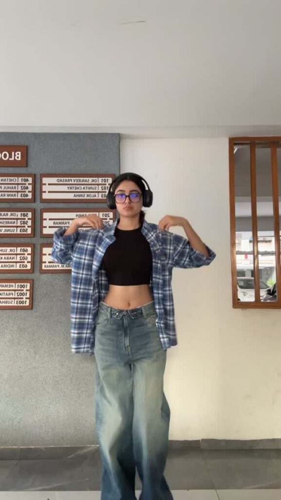 A young woman posing in an open blue plaid flannel shirt and wide-leg denim jeans.