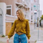 Woman smiling while wearing a mustard yellow knit jumper and blue denim jeans on a city street.