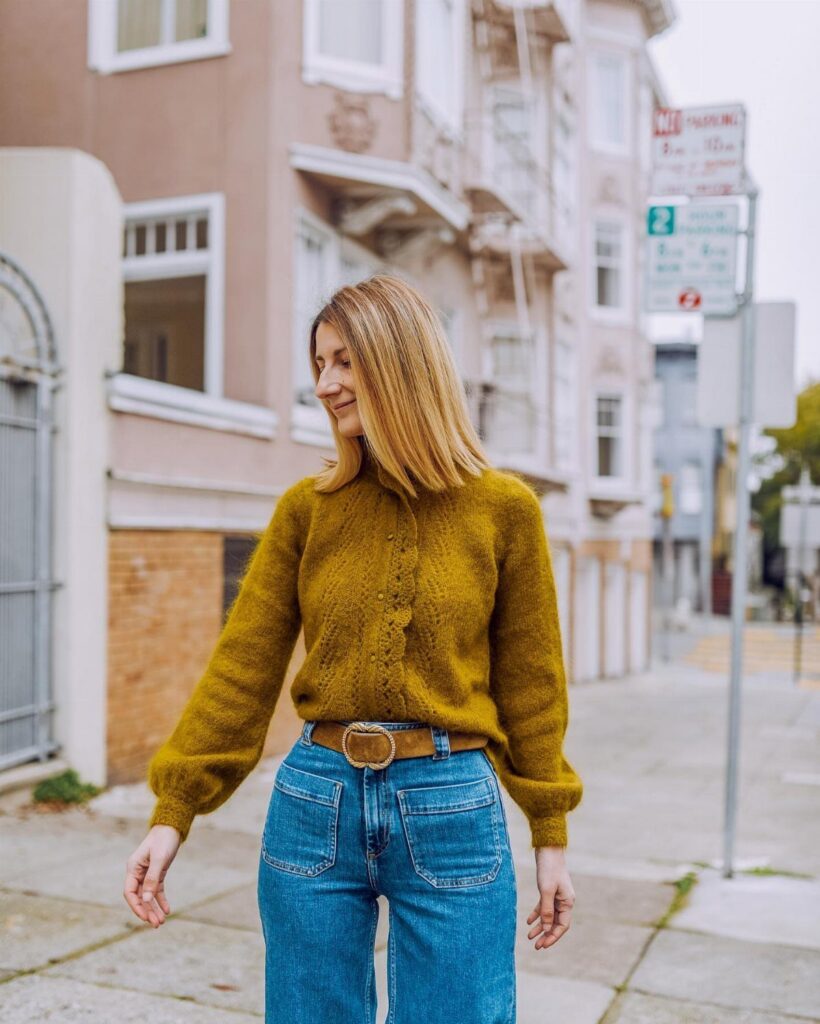 Woman smiling while wearing a mustard yellow knit jumper and blue denim jeans on a city street.