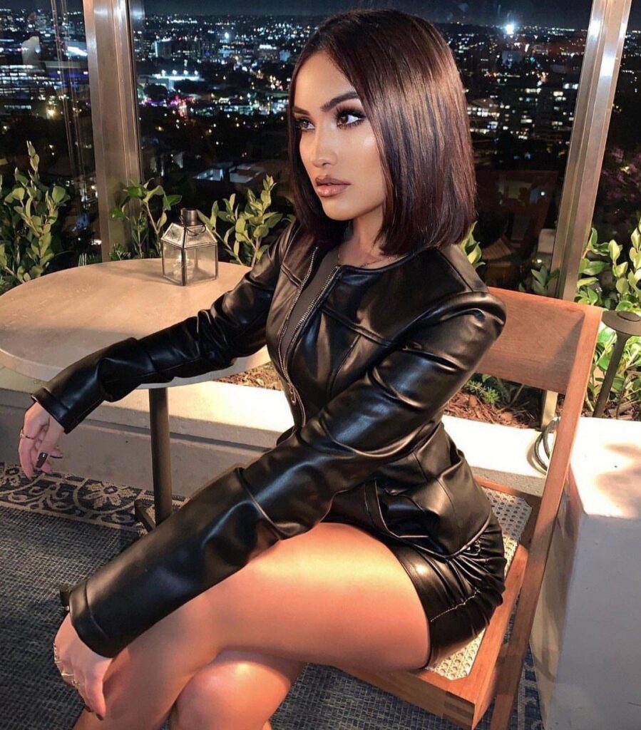 A woman wearing a black leather jacket dress sits at a table overlooking a city skyline.
