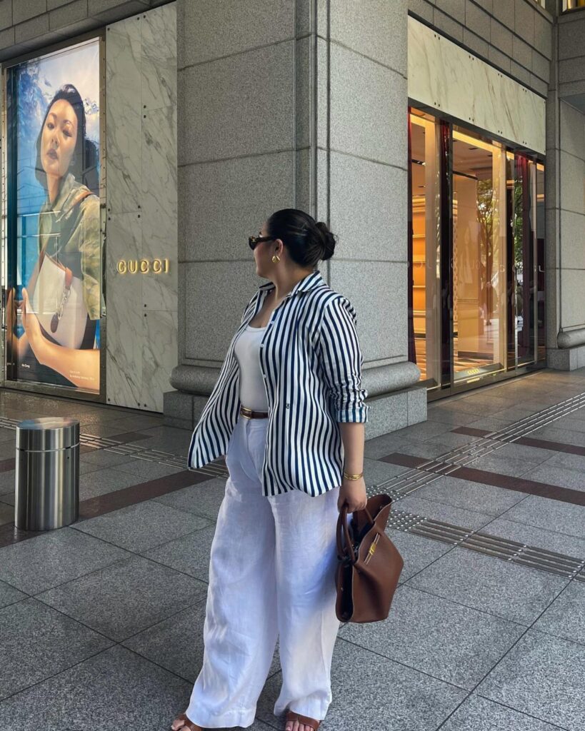 A woman in a striped shirt and white wide-leg trousers walking on a city street.