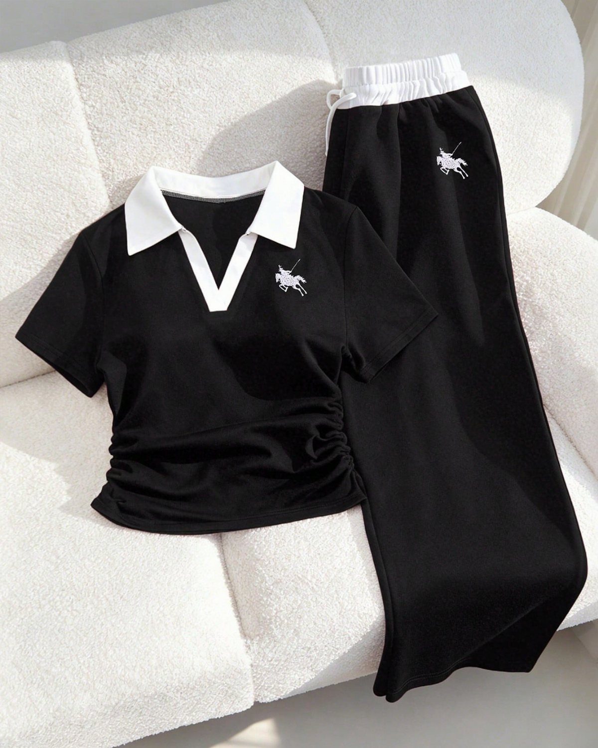 Black polo shirt and matching drawstring pants with white collar and emblem on a white sofa.