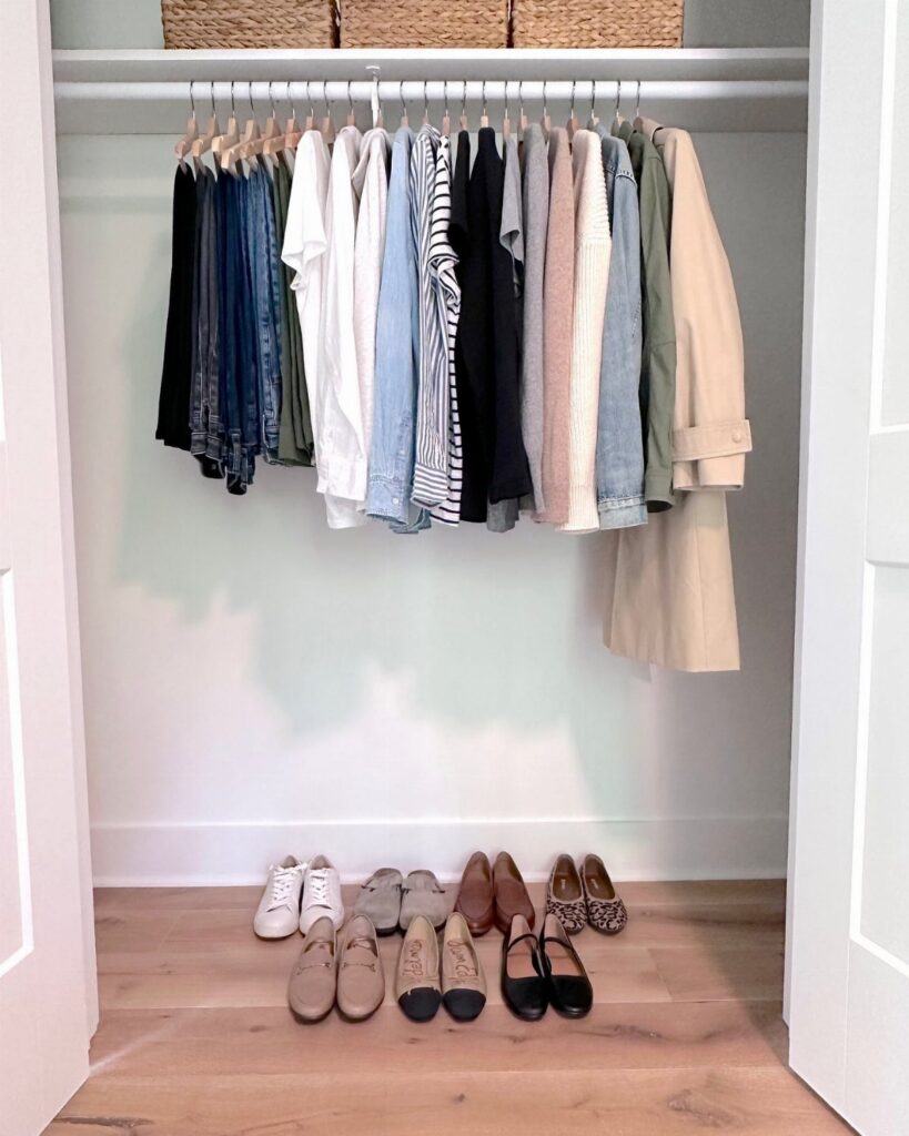 Various neutral clothing items hanging in an open closet with shoes lined up on the floor below.