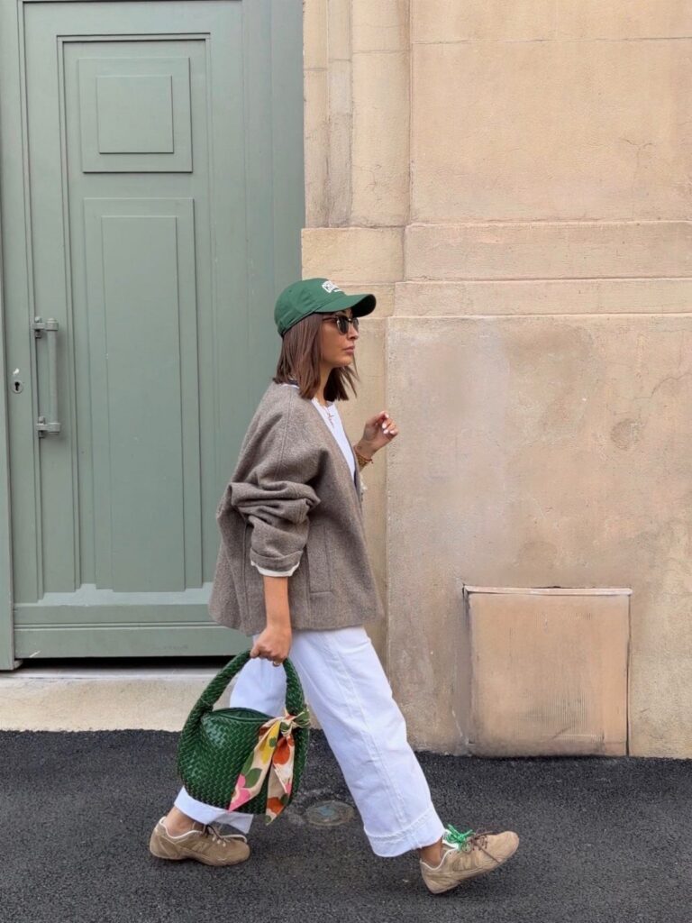 A woman walking in a brown sweater, white trousers, and a green cap while carrying a green handbag.