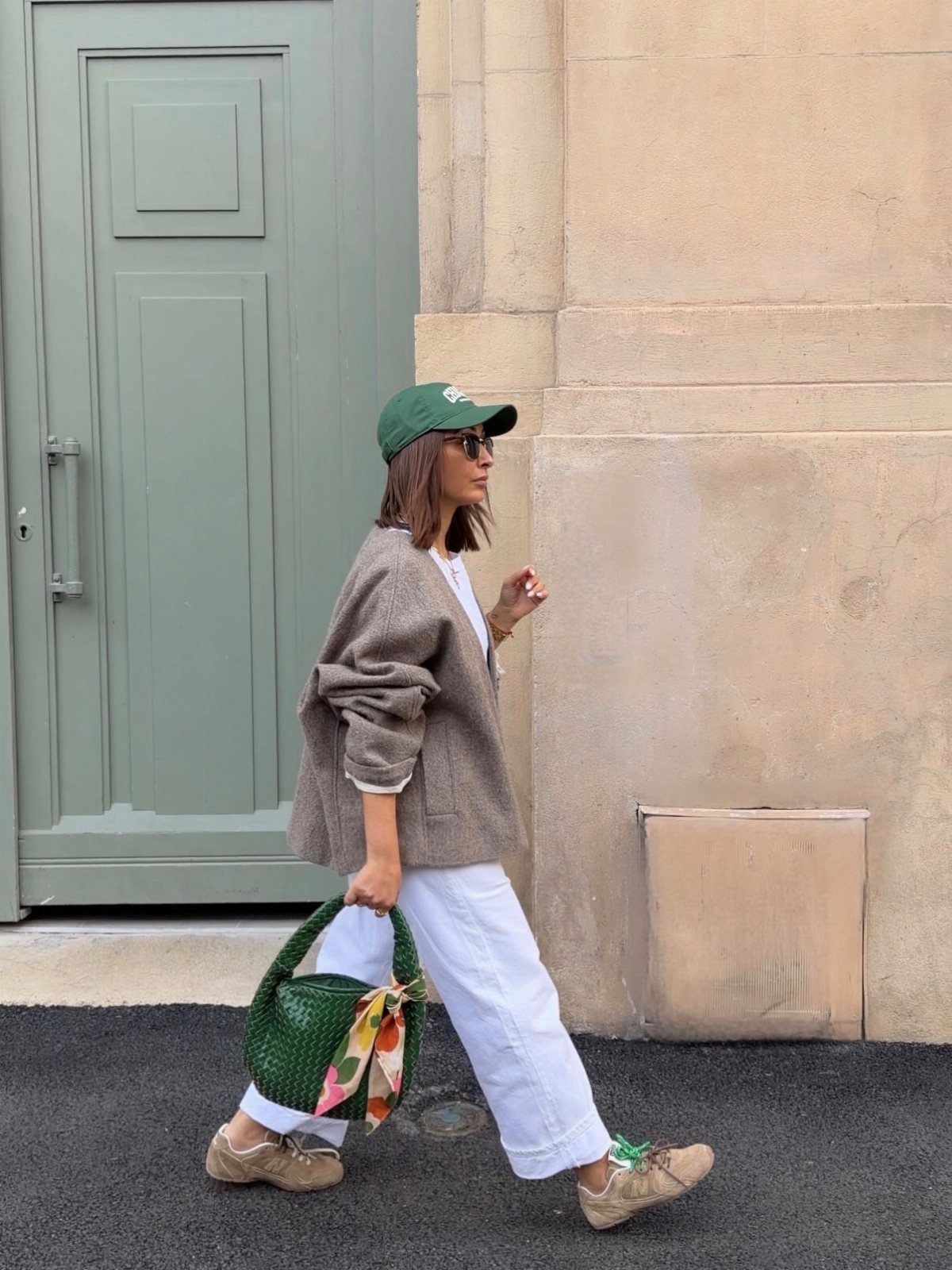 A woman walking in a brown sweater, white trousers, and a green cap while carrying a green handbag.