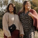 Two women smiling outdoors wearing autumn fashion including a beige sweater and a floral dress.