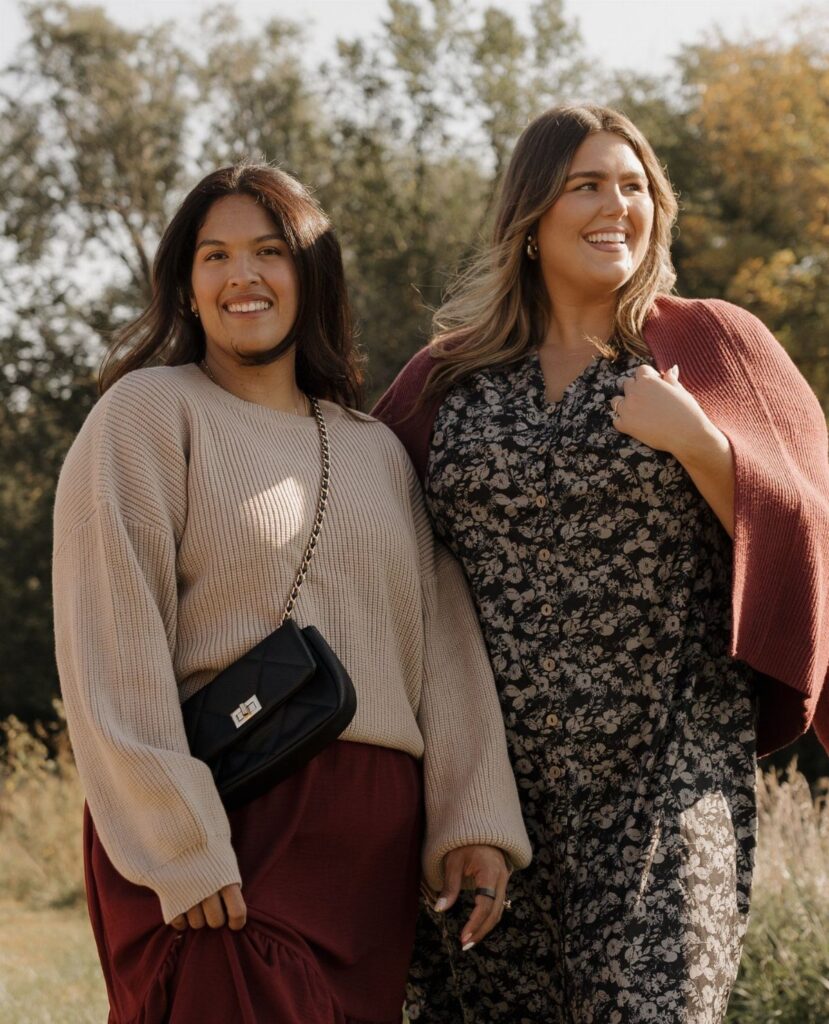 Two women smiling outdoors wearing autumn fashion including a beige sweater and a floral dress.