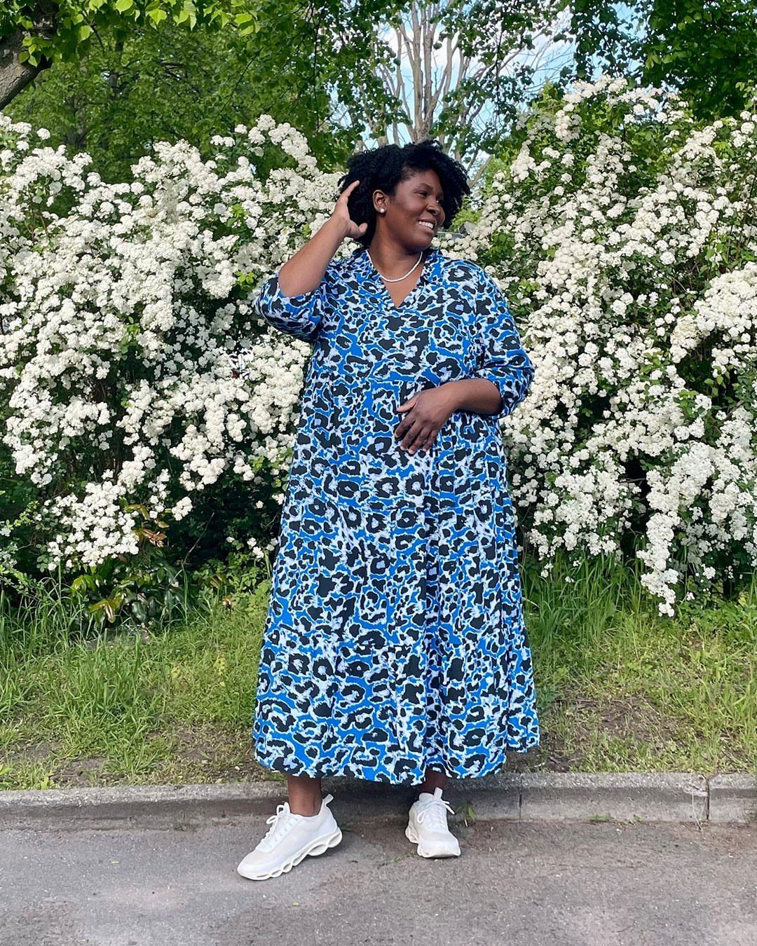 A woman wearing a blue leopard print midi dress and white sneakers standing by bushes.