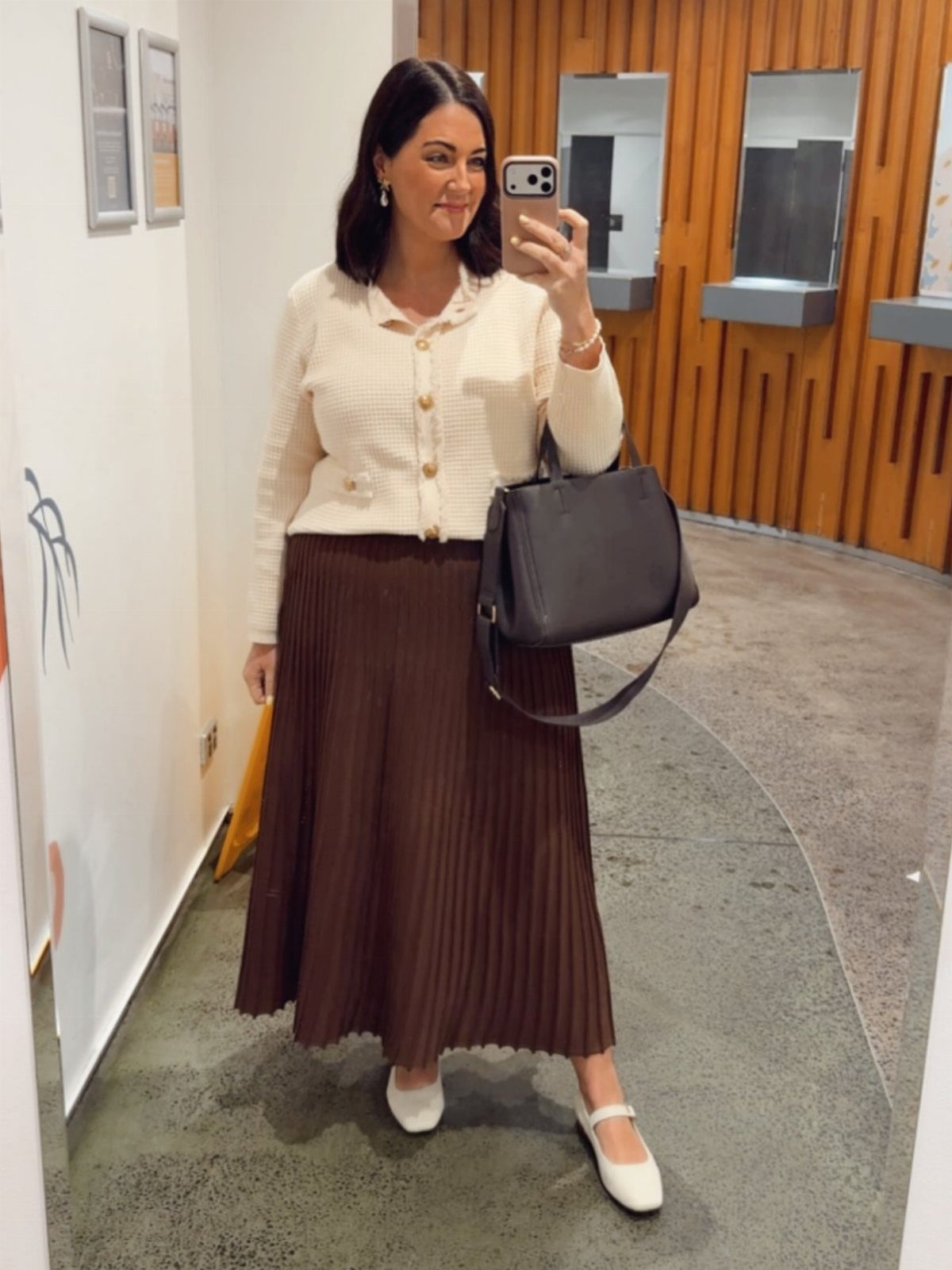 A woman posing in a cream cardigan and brown pleated skirt for a mirror selfie.