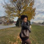 A woman in a leather jacket and black tights poses in a park with autumn leaves.