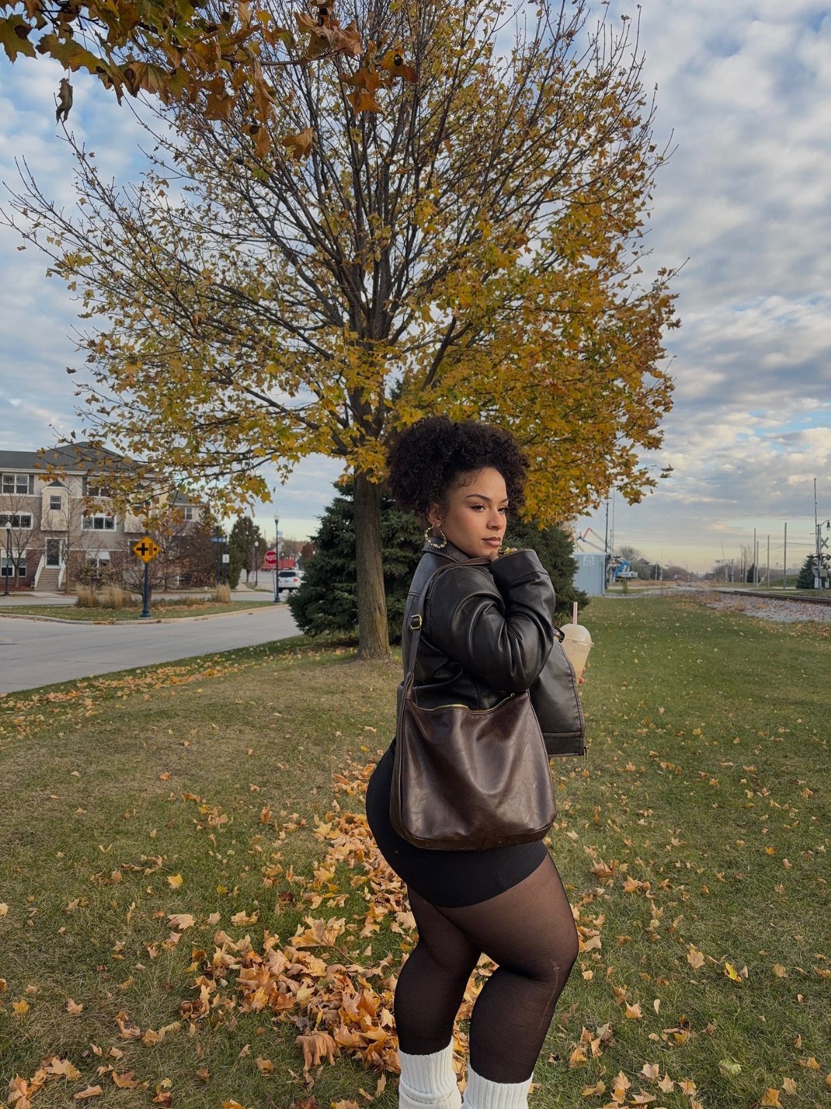 A woman in a leather jacket and black tights poses in a park with autumn leaves.