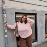 Woman in a pink oversized knit sweater and brown trousers posing on a city street.