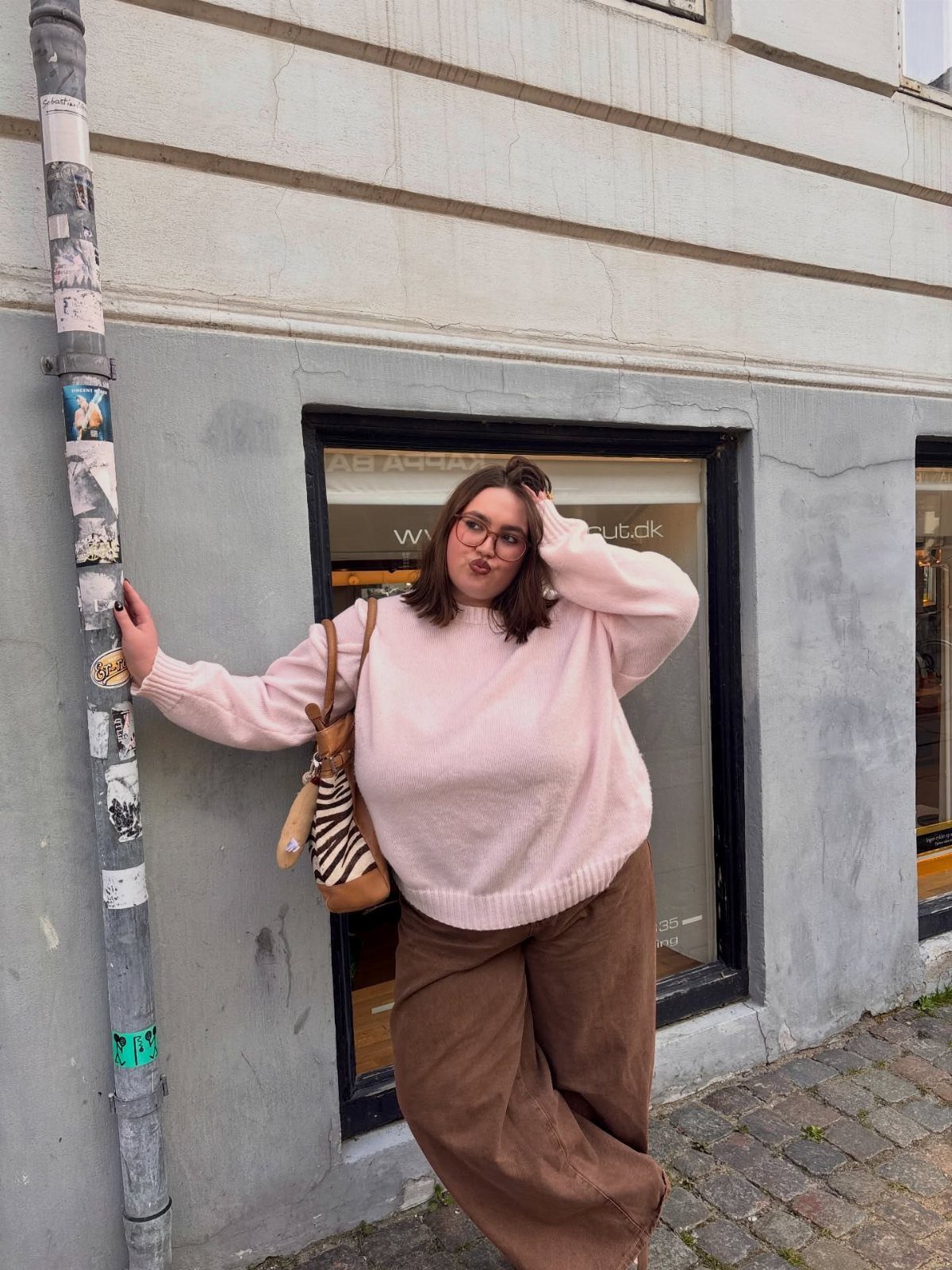 Woman in a pink oversized knit sweater and brown trousers posing on a city street.