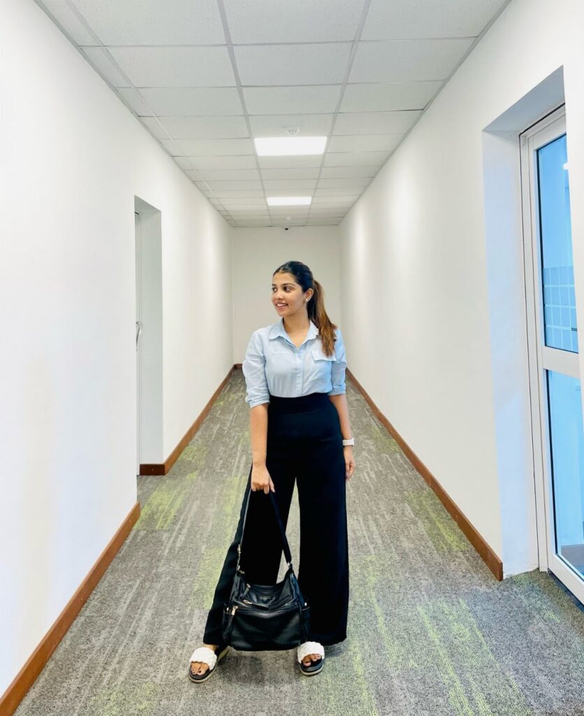 A woman in a light blue shirt and black trousers stands in an office hallway holding a handbag.