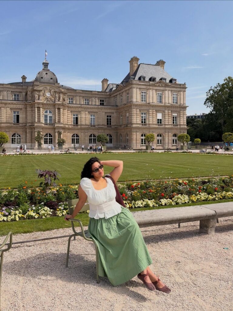 A woman in a white blouse and long green skirt sitting on a chair in a park.