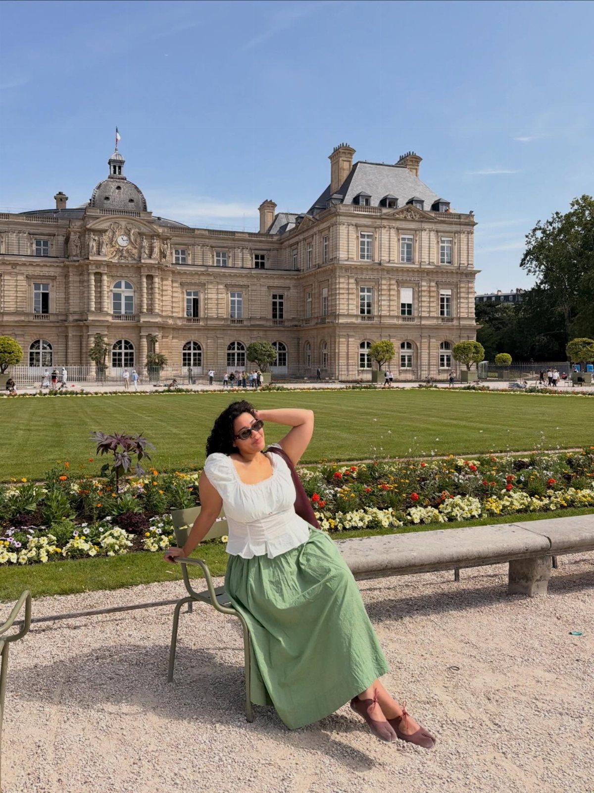 A woman in a white blouse and long green skirt sitting on a chair in a park.