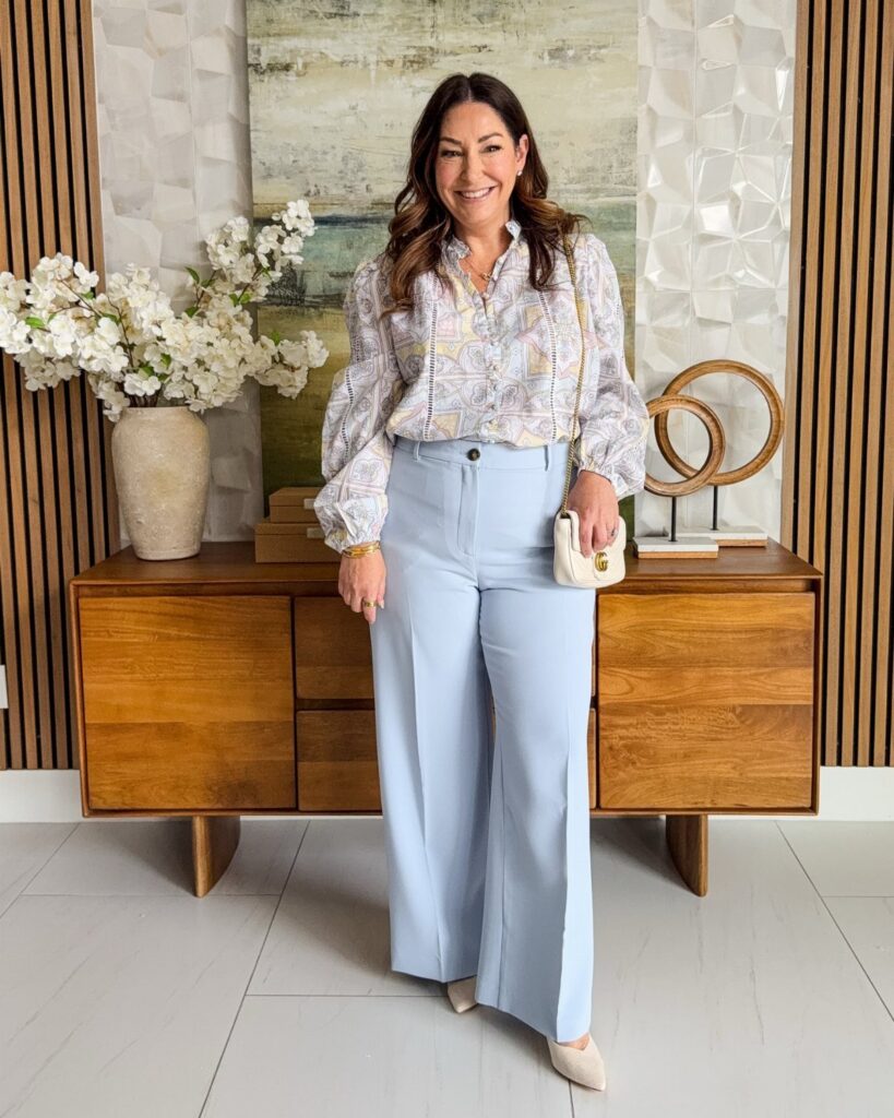 A woman posing in a patterned blouse and light blue wide-leg trousers with a small white handbag.