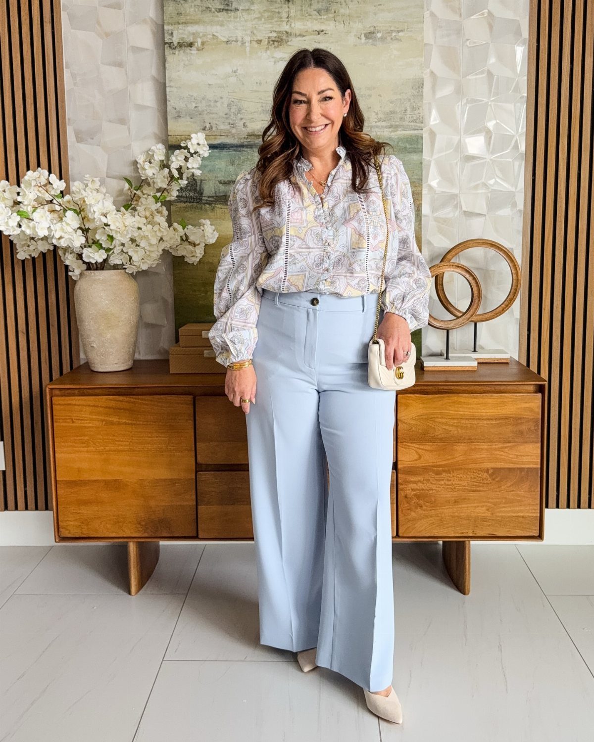 A woman posing in a patterned blouse and light blue wide-leg trousers with a small white handbag.