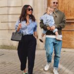 A woman wearing a striped shirt and black trousers walks next to a man holding a baby.