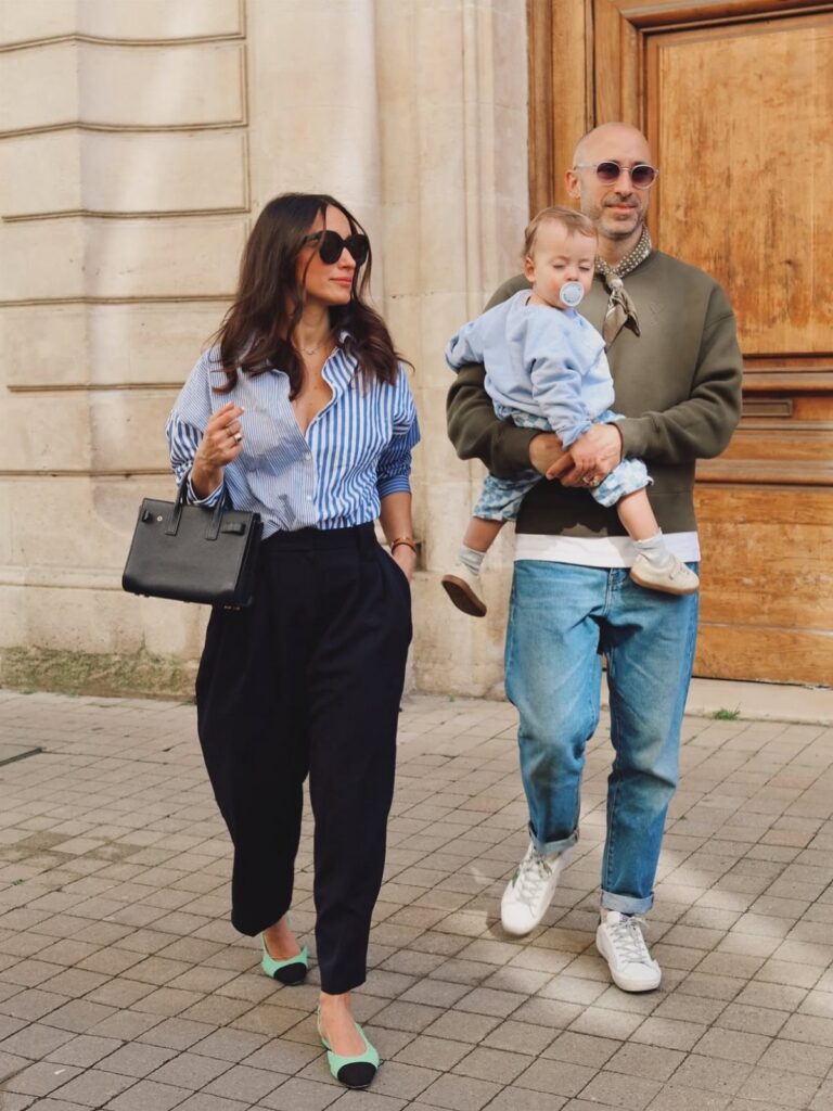 A woman wearing a striped shirt and black trousers walks next to a man holding a baby.