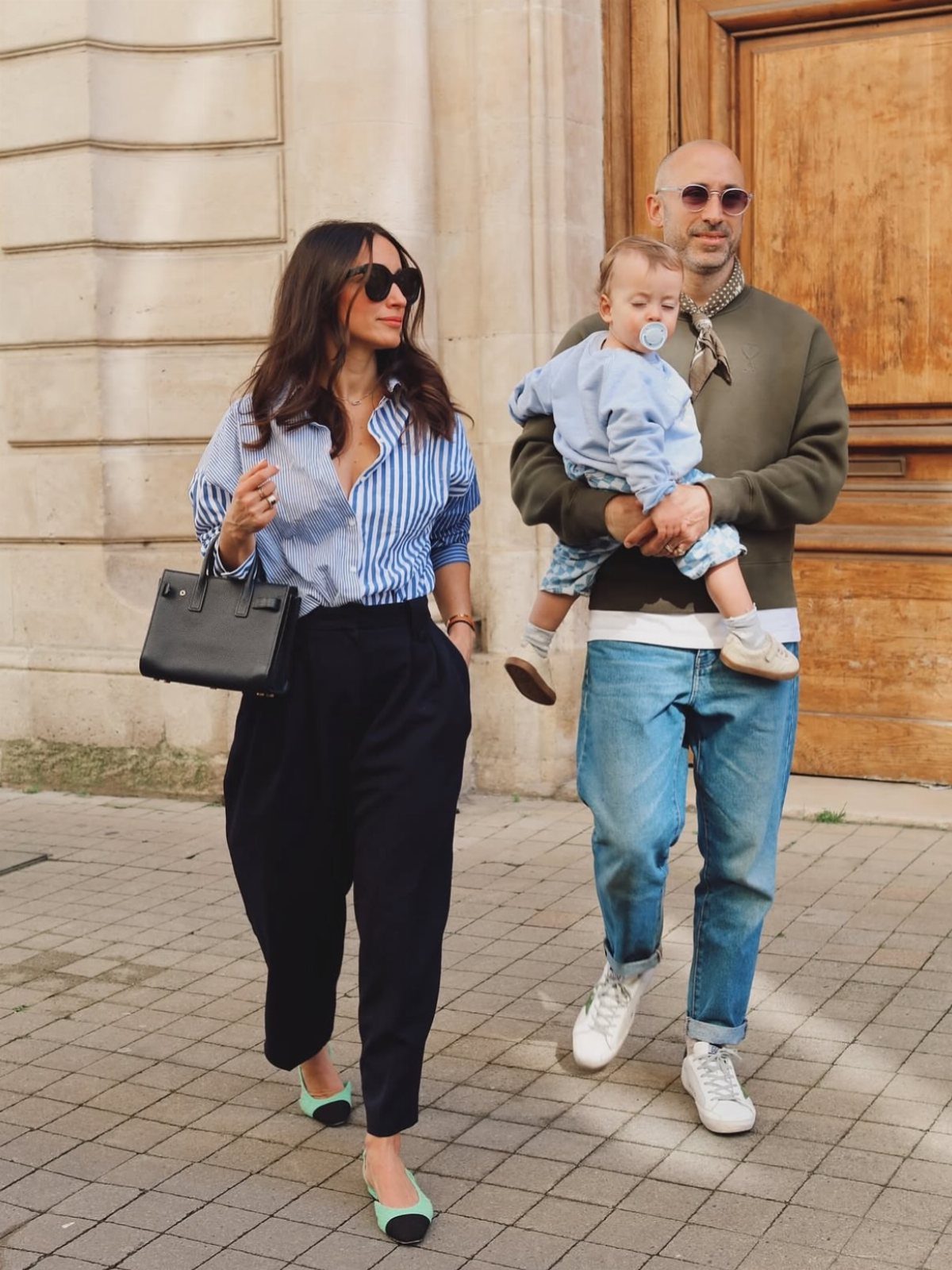 A woman wearing a striped shirt and black trousers walks next to a man holding a baby.