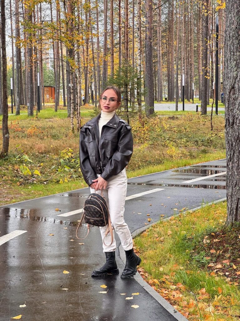 Woman wearing white jeans and a black leather jacket standing on a wet forest path.