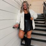A woman posing on stairs wearing a white jacket, black skirt, and black boots.