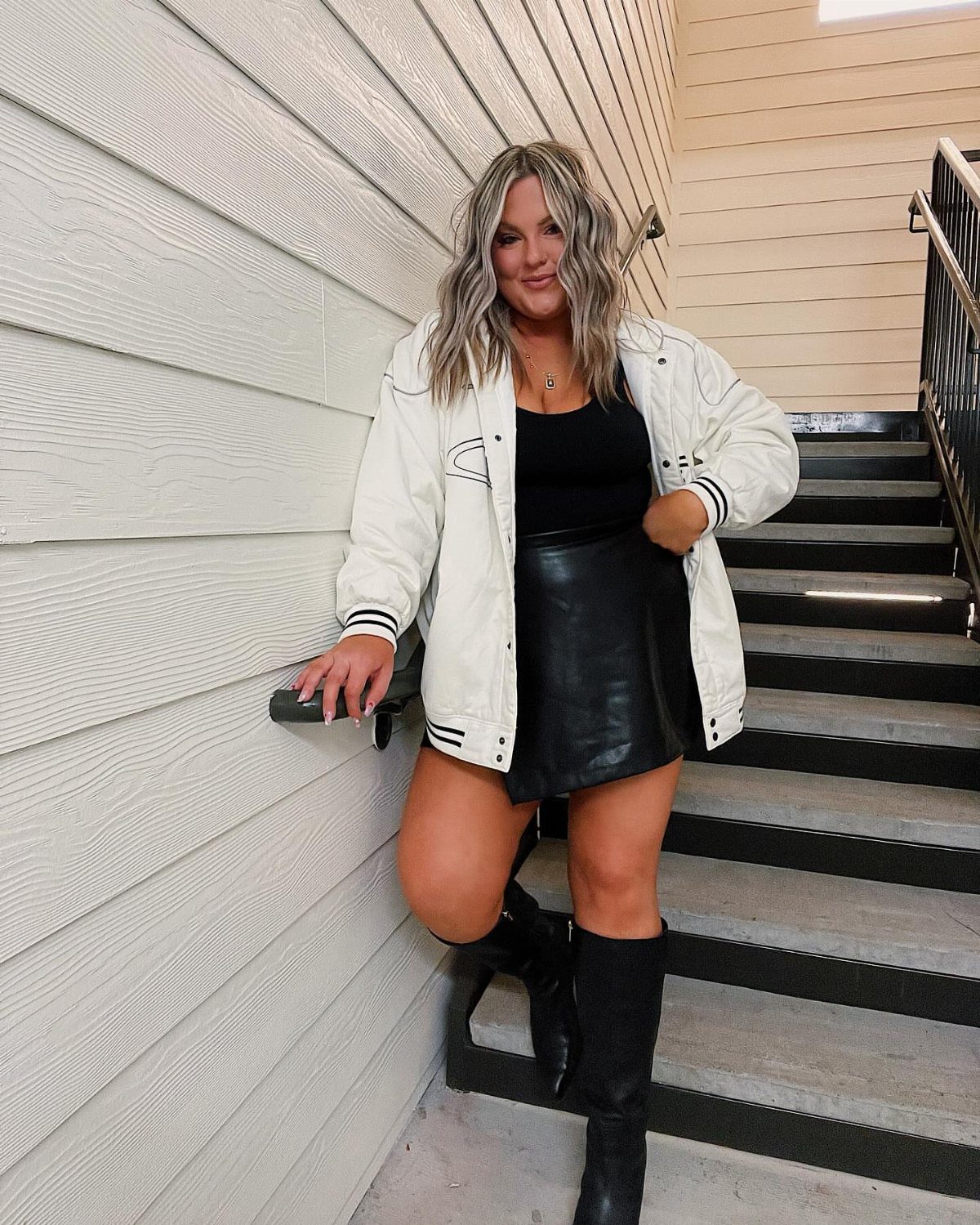 A woman posing on stairs wearing a white jacket, black skirt, and black boots.